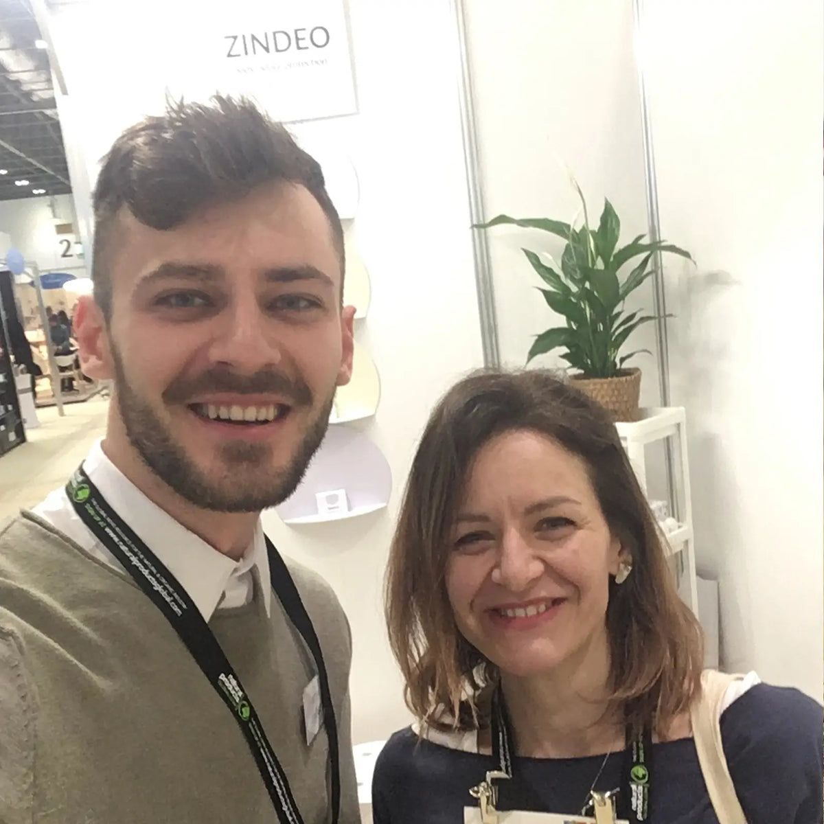 Two smiling people at a trade show booth with a ZINDEO sign and a plant behind them