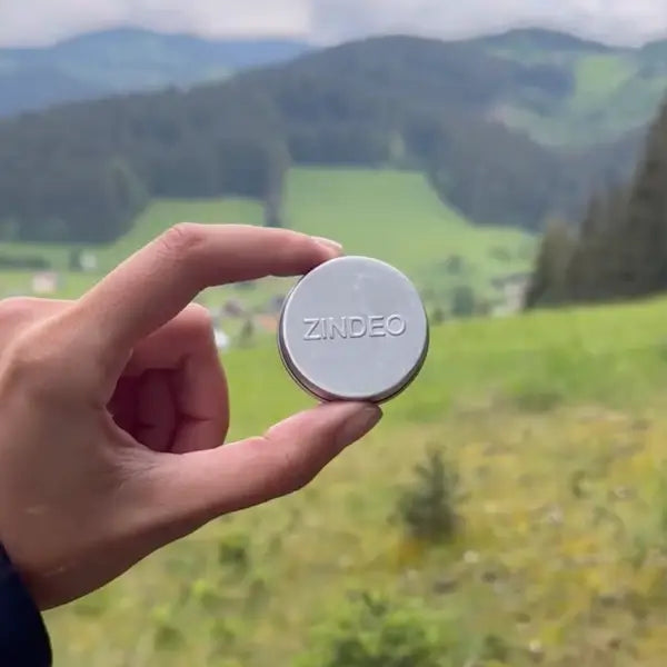 A hand holding a round silver ZINDEO deodorant tin against green mountains and fields