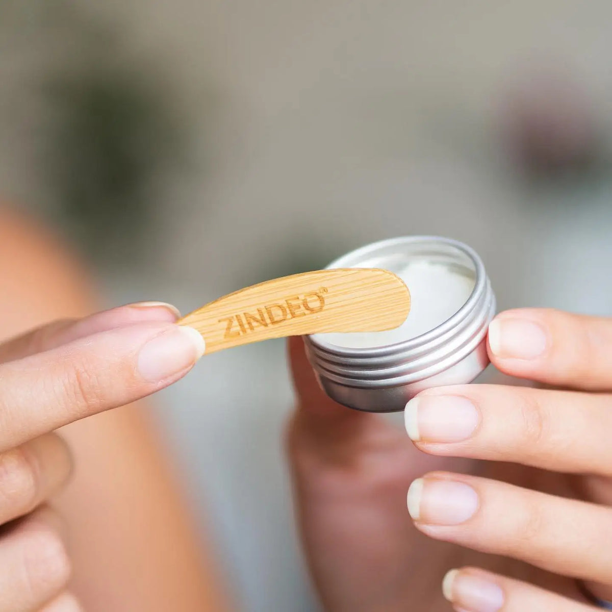 Hands holding an open tin of deodorant cream with a wooden ZINDEO applicator spatula