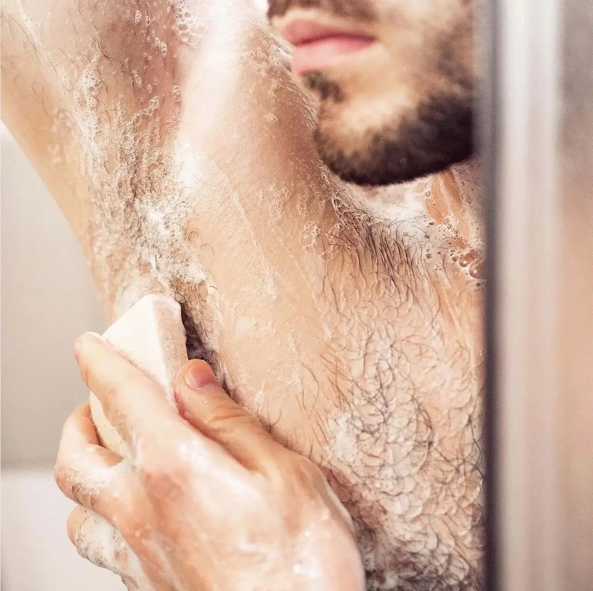 A bearded man in the shower washing his underarm with a lathered soap bar